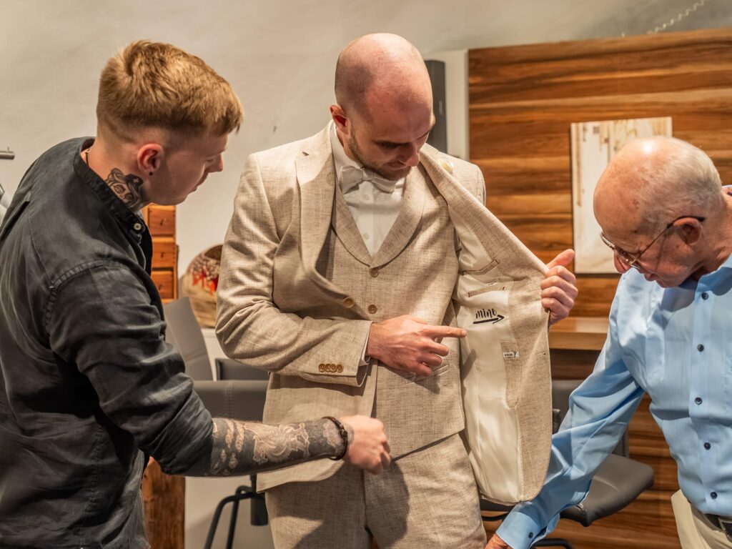 Mann probiert Hochzeitsanzug an, sein Freund und ein älterer Herr helfen ihm